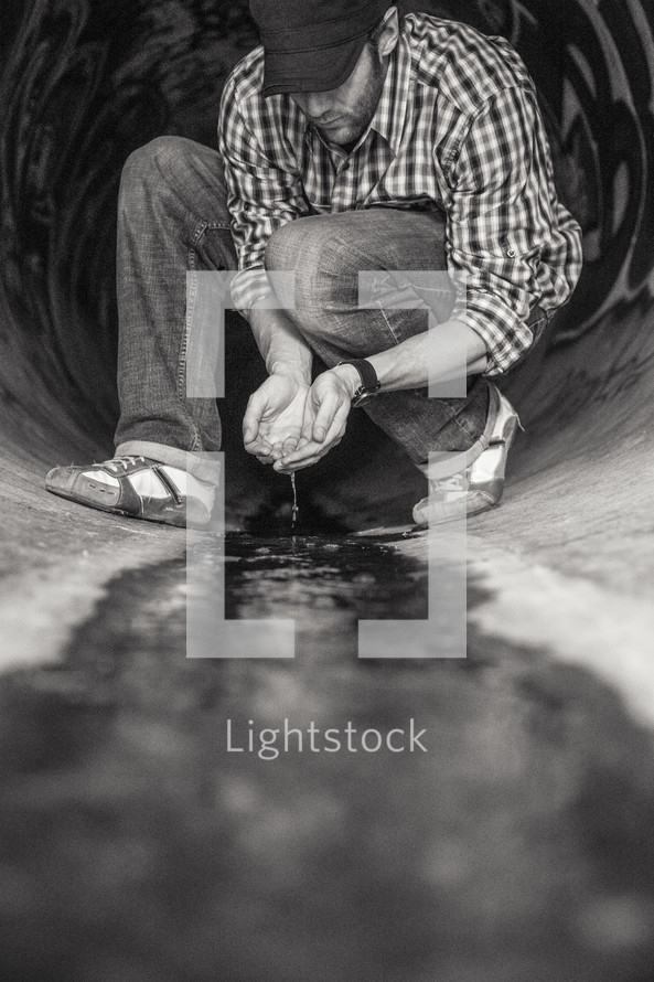 Man cupping flowing water with his hands while kneeling in a sewer drain pipe painted with graffiti.