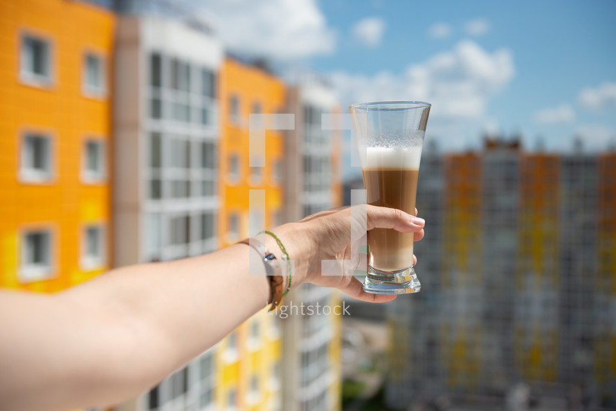 multilayer coffee or cappuccino in a glass of coffee on a woman's hand stretched out into the window against the background of an apartment building