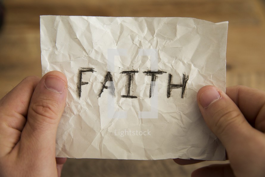 hands holding a piece of crumpled paper with the word faith