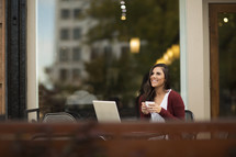 a young woman sitting at an outdoor table with on a laptop and drinking coffee 