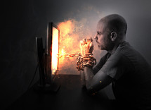 A man is hopelessly chained to a burning computer screen