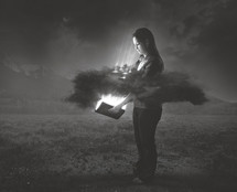A woman holds a bright glowing Bible that shines through a dark cloud around her