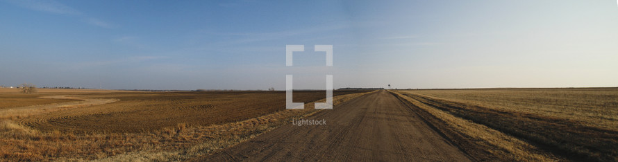Panorama of a field and dirt road in the middle of nowhere Kansas with a blue sky. 