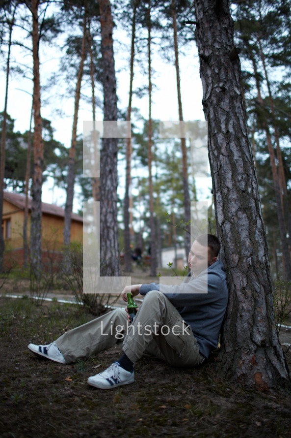 Young sorrow man sitting near the pine tree, drinks beer and thinking about life