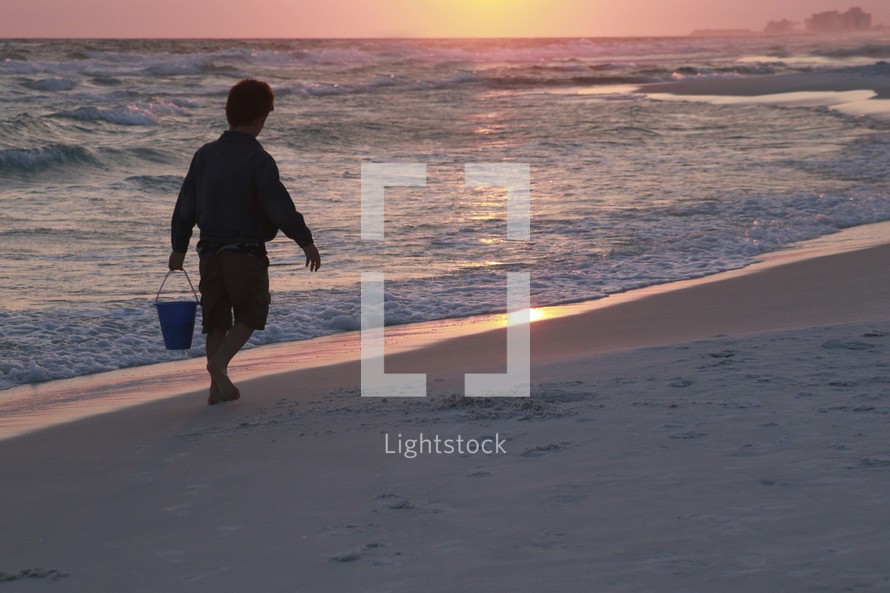 a boy carrying a sand bucket on a beach at sunset 