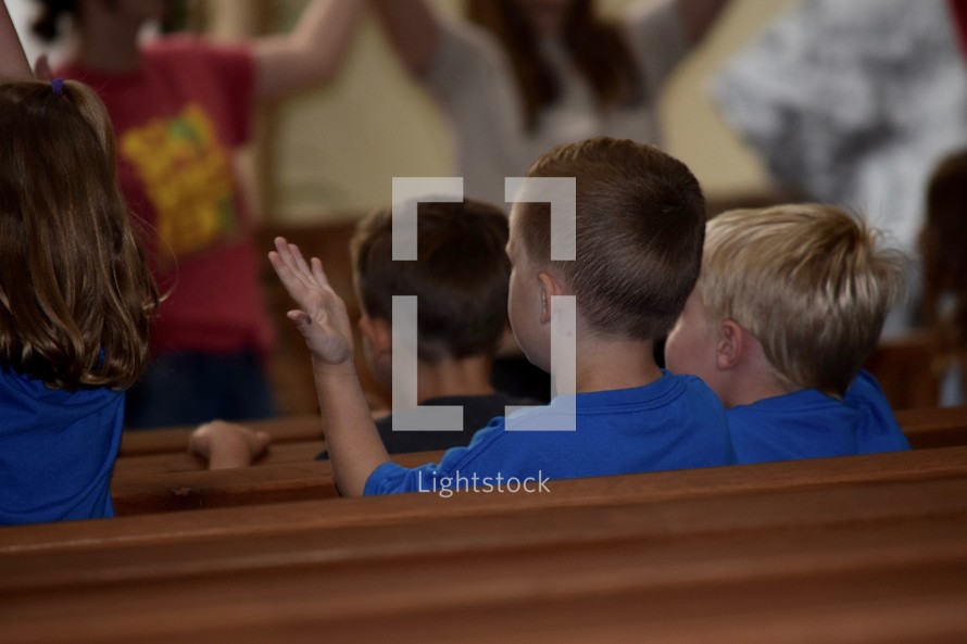 kids with raised hands sitting in church pews singing songs at VBS 