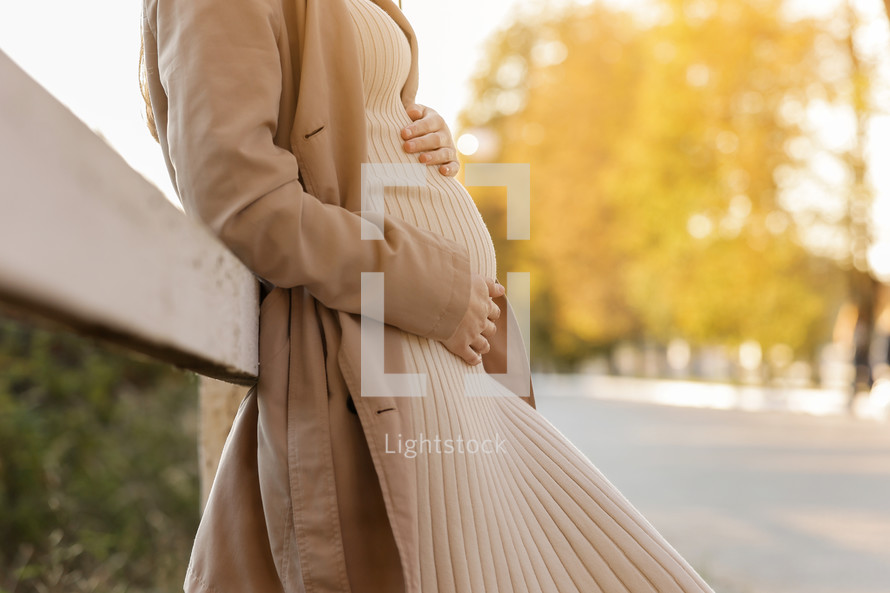 Cropped photo of pregnant woman holds hands on belly in the autumn park. Young woman in maternity warm dress and coat waiting for baby birth. Pregnancy, motherhood, mothers day. Happy pregnancy.