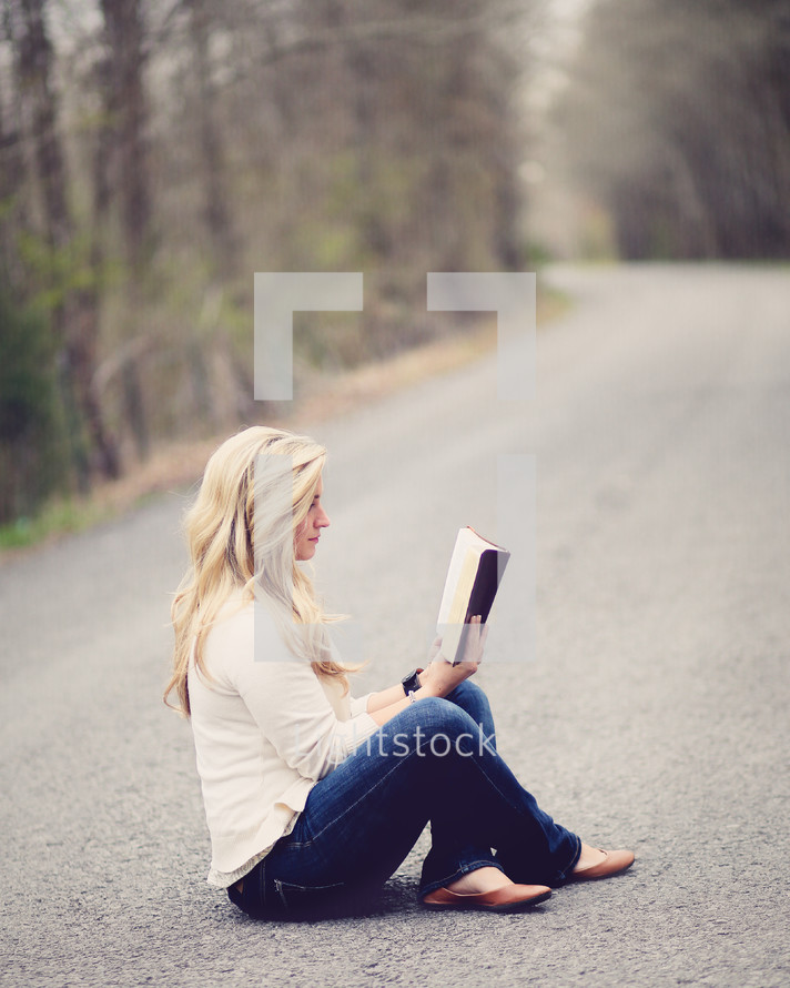 Woman sitting with legs crossed in the middle of the road reading the Bible.