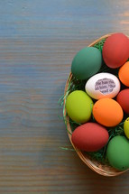 a white eggshell with a piece of the bible inside saying: HE HAS RISEN! between colorful painted eggs in a basket 