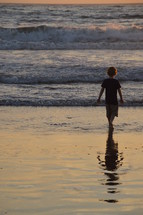 A child wading in the water on the beach at sunset.