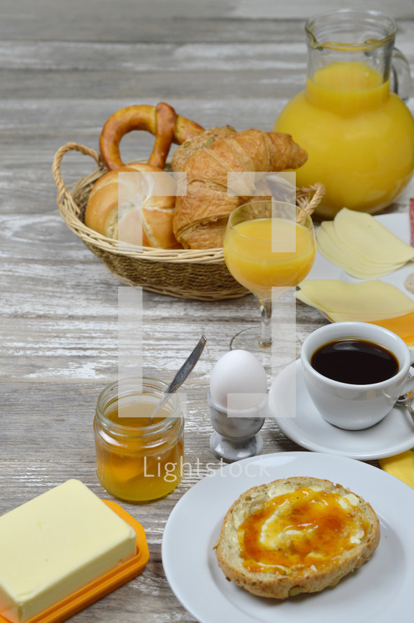 breakfast table with lots of fresh food like coffee, rolls, cheese, sausages, eggs, orange juice, jam, butter and a basket full of croissant, rolls and pretzel with a little copy space above