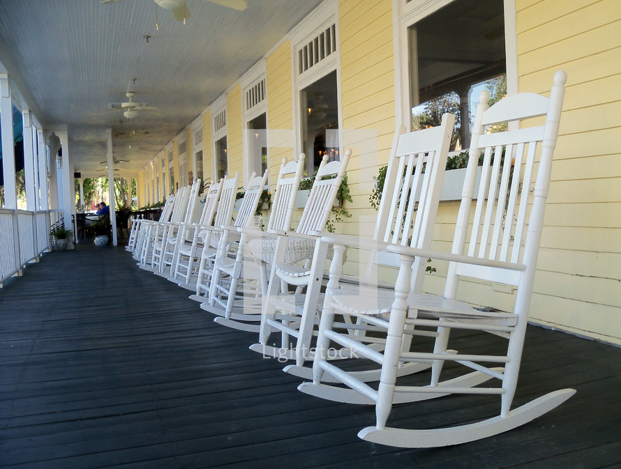 A line of white rocking chairs on a long wooden front porch in a rural country setting. 