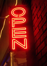 A Neon Open sign lights up the night in neon orange surrounded by Christmas lights illuminating a brick exterior building in a downtown retail area telling passer-bys that the store is open for business and welcoming the public during the shopping retain season.. 