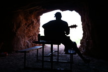 silhouette of young man sitting on a picnic table in a cave on a beach playing a guitar 