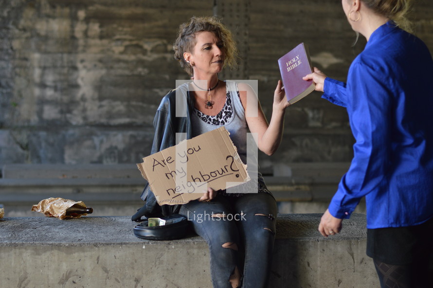 a woman handing a Bible to a homeless woman begging 