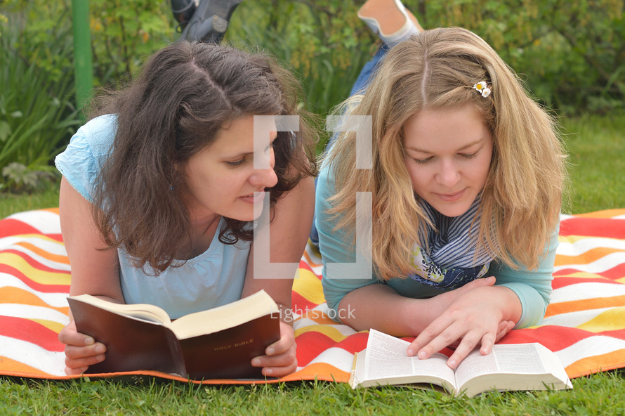 Young women smiling while reading in the bible together laying outside on a blanket in the grass on a sunny summer day. 
