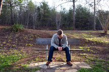 man with head bowed and praying hands sitting on a bench outdoors