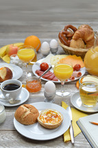 breakfast table with lots of fresh food like coffee, rolls, cheese, eggs, orange juice, tea, jam, butter, banana, apple, orange and a basket full of croissant, rolls and pretzel with copy space above