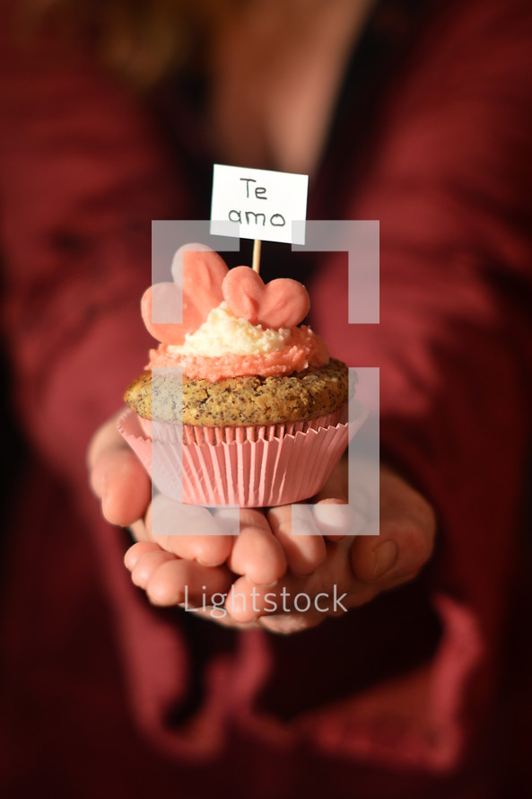 Woman offering a cupcake with pink hearts and a sign saying TE AMO.

