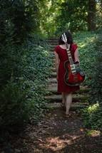 a woman in a red dress with an electric guitar walking through the dark woods