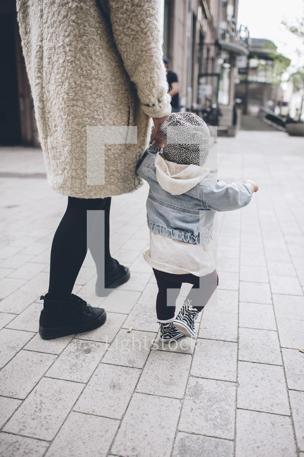 mother and toddler daughter walking holding hands on a sidewalk 