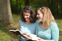 Young women smiling while reading in the bible together sitting outside on a bench on a sunny day. 