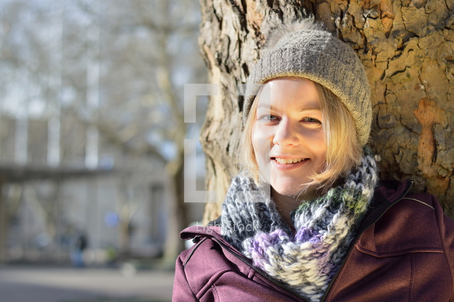 smiling woman in a beanie leaning against a tree 