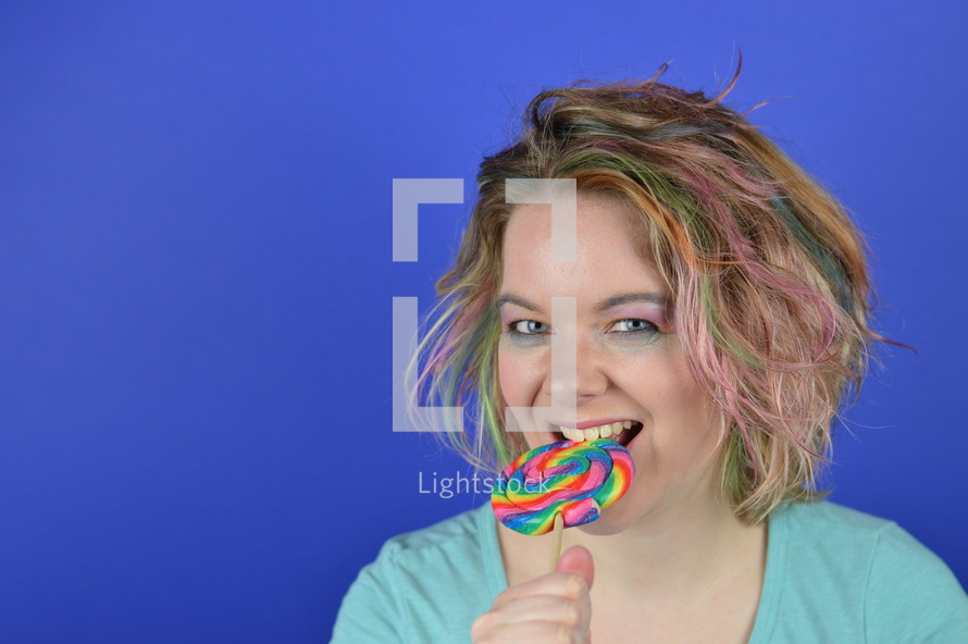 portrait of a young blond woman with colourful streaks in the hair biting into a big sweet multicolored lollipop and having fun looking into the camera