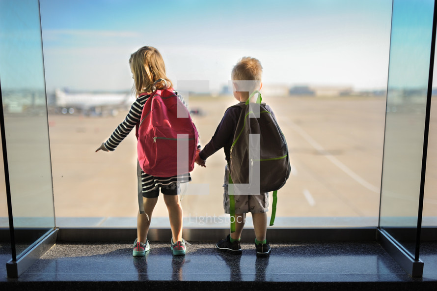 siblings holding hands looking out a window at an airport 