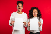 Portrait of young handsome african american man and woman couple holding white empty paper blanks on red studio background. Copy space. Match with separate sign