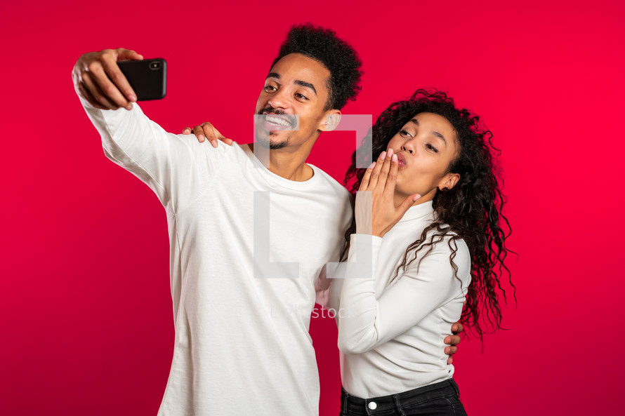 Young african american couple in white making selfie with smartphone on red studio background.Lovers match, love, holidays, happiness concept.