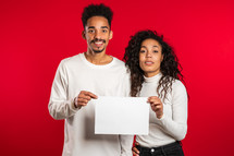 Portrait of young handsome african american man and woman couple holding white empty paper blanks on red studio background. Copy space. Match with separate sign