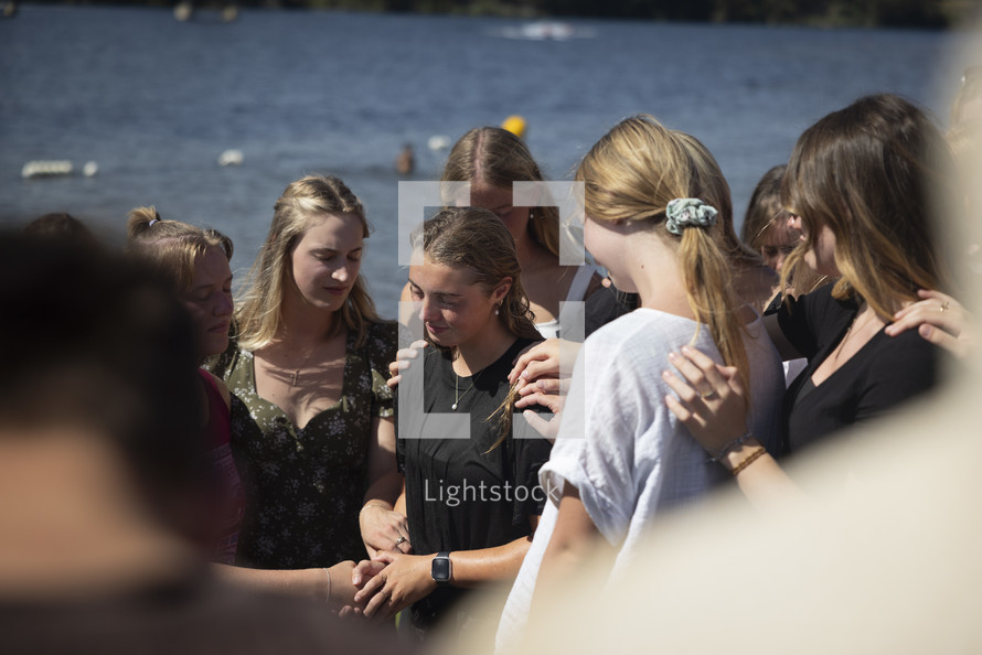 people surrounding and praying for a young woman who has been baptized by a lake 