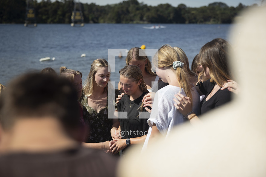 people surrounding and praying for a young woman who has been baptized by a lake 