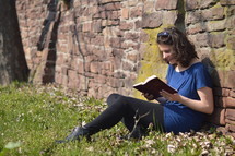 Woman reading the bible while sitting in a meadow next to a stone wall.