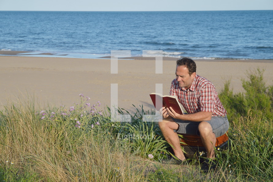 a man reading a Bible sitting on suitcase on a beach 
