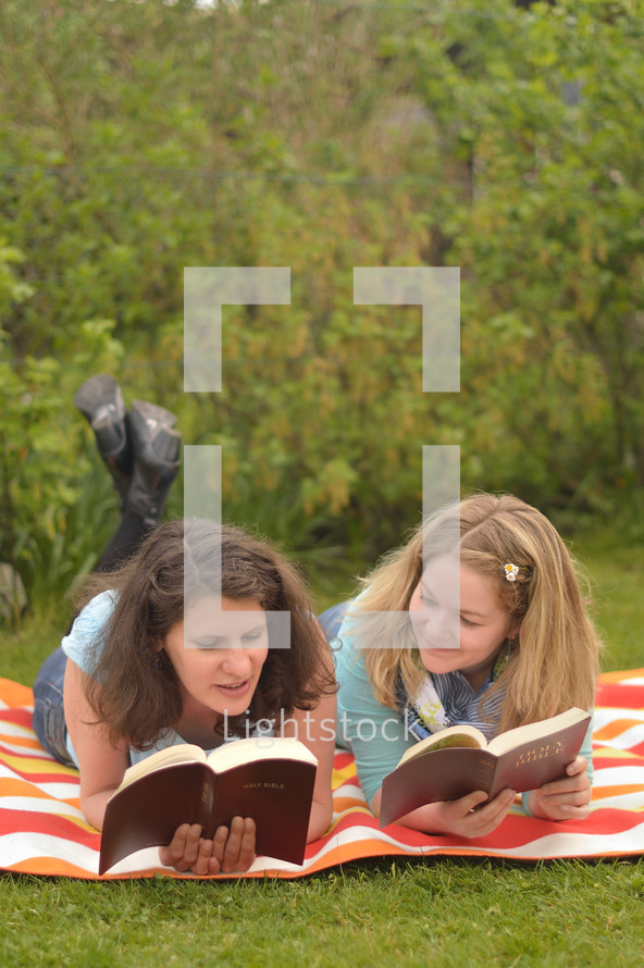 Young women smiling while reading in the bible together laying outside on a blanket in the grass on a sunny summer day. 
