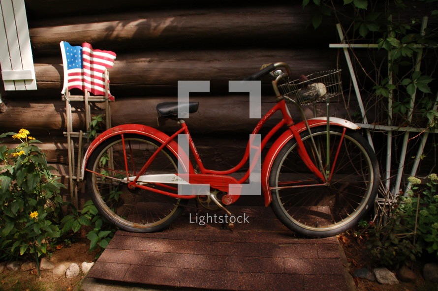 An American flag and a red bicycle leaning against a log building.