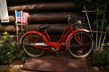 An American flag and a red bicycle leaning against a log building.