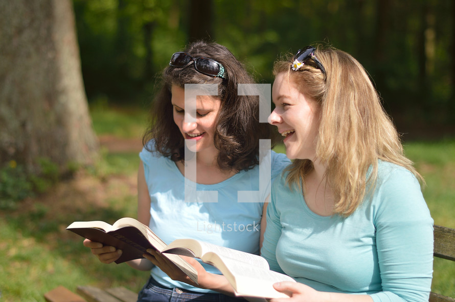 Young women smiling while reading in the bible together sitting outside on a bench on a sunny day. 
