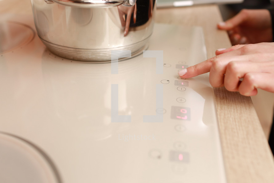 Female hand turns on electric hob closeup, finger presses sensor button on modern white induction cooker. Metal steel saucepan on kitchen stove top panel, modes of cooking surface, Domestic equipment.