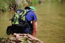 man with a backpack squatting near a river 