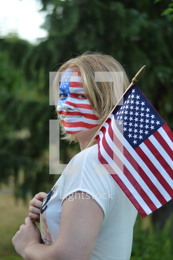 woman with American flag face paint - young blond woman with the american flag painted all over her face carrying a flag over her shoulder