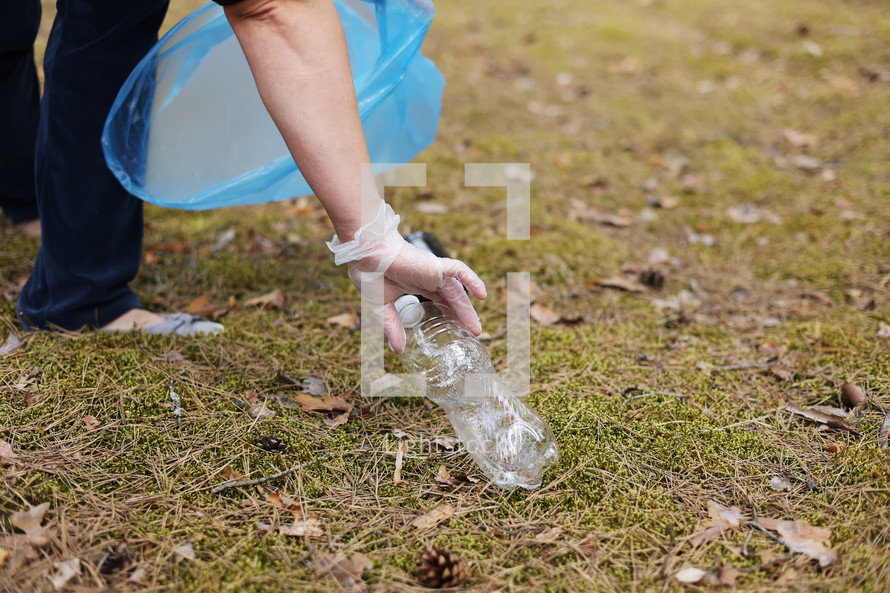 A woman hands in gloves collects and puts used plastic bottle into a blue trash bag. A volunteer cleans up the park on a sunny bright day. Clearing, pollution, ecology and plastic concept.