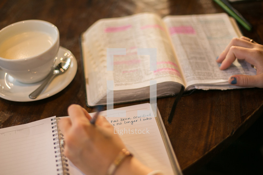 a woman reading a Bible and writing in a journal sitting at a table in a coffee shop 