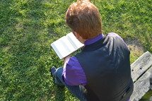man reading a Bible sitting on a park bench 