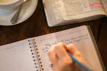 a woman reading a Bible and writing in a journal sitting at a table in a coffee shop 
