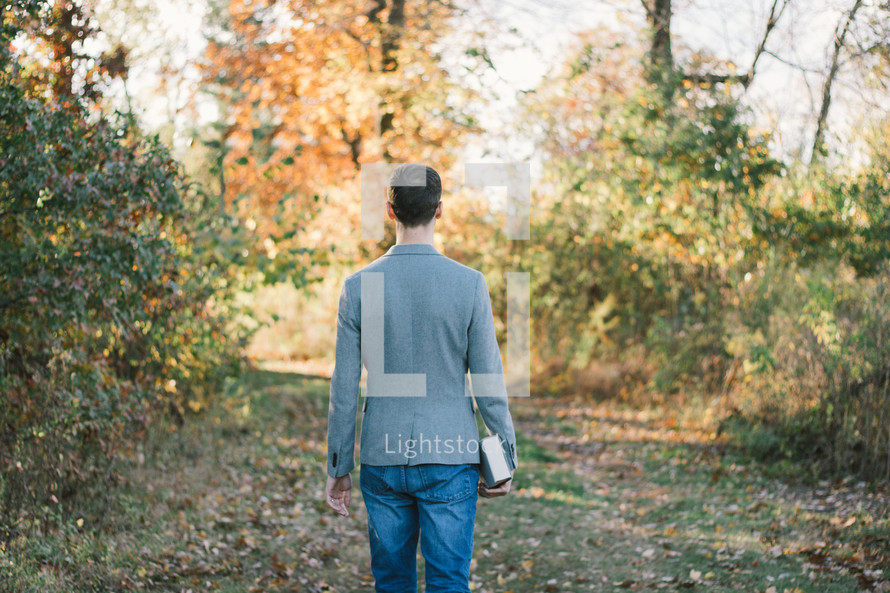 a man in a blazer carrying a Bible walking into a fall forest 