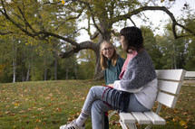 Two young women sitting and talking, smiling on a bench with lovely background
