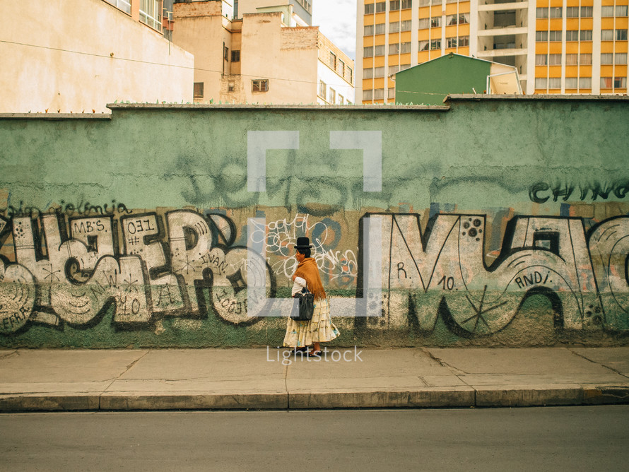 A Latin American woman walks along a graffiti covered wall.